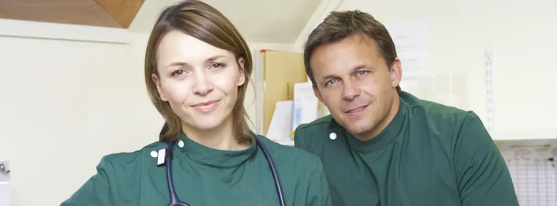 Un vétérinaire et sa conjointe en blouse verte, stéthoscope autour du cou, dans la clinique